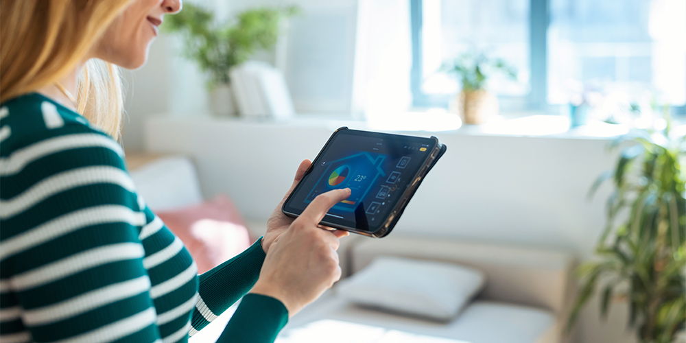 Eine Frau bedient ein Tablet in einem hellen Wohnraum mit Pflanzen. Auf dem Bildschirm ist eine Steuerungsoberfläche zu sehen.