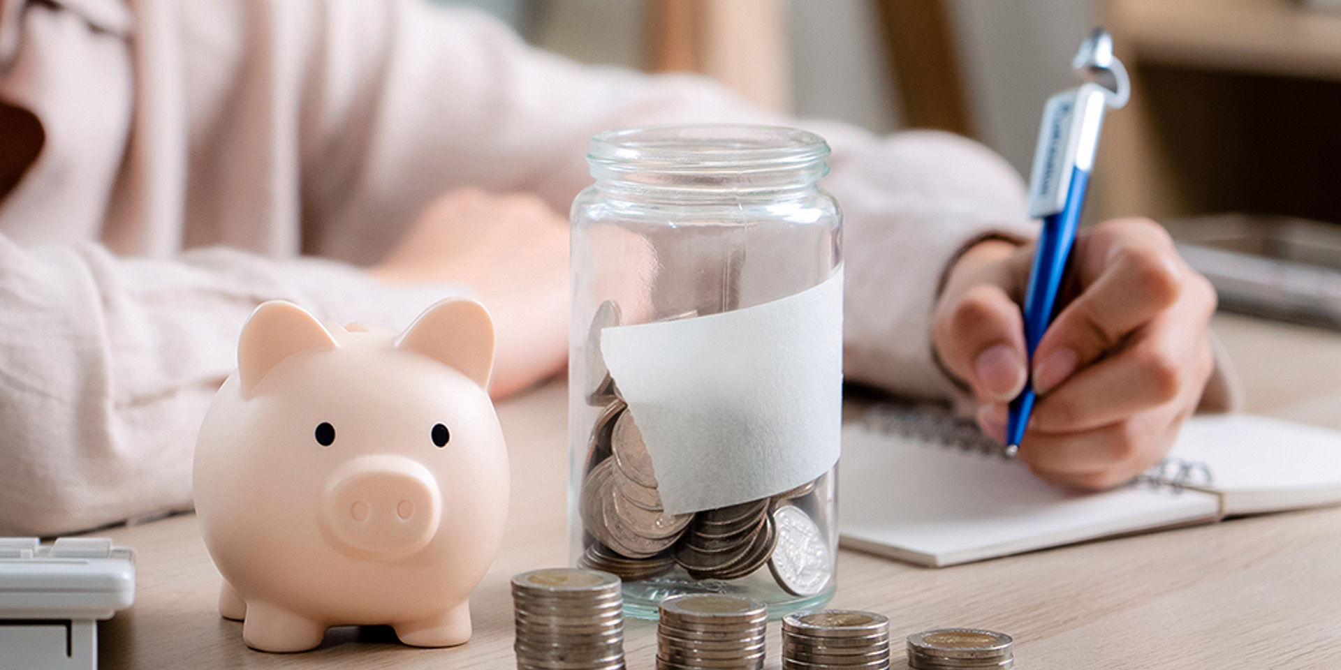 Ein Sparschwein und ein Glas mit Münzen stehen auf einem Tisch, daneben werden Notizen gemacht. Die Szene steht für Sparen und Finanzplanung.