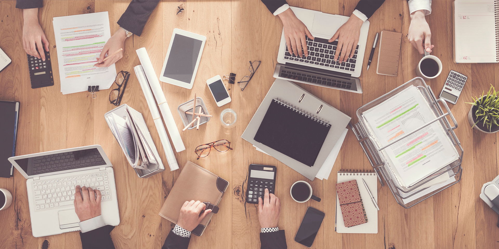 Mehrere Personen arbeiten an einem Tisch mit Laptops, Unterlagen und Taschenrechnern. Die Aufnahme zeigt eine organisierte Bürosituation aus der Vogelperspektive.