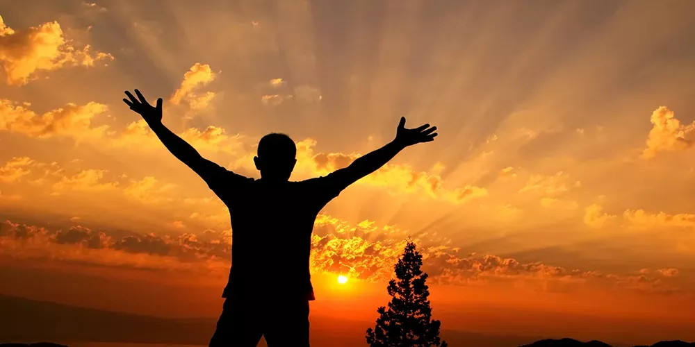 Die Silhouette eines Mannes vor einem Sonnenaufgang, der mit weit geöffneten Armen den neuen Tag begrüßt.