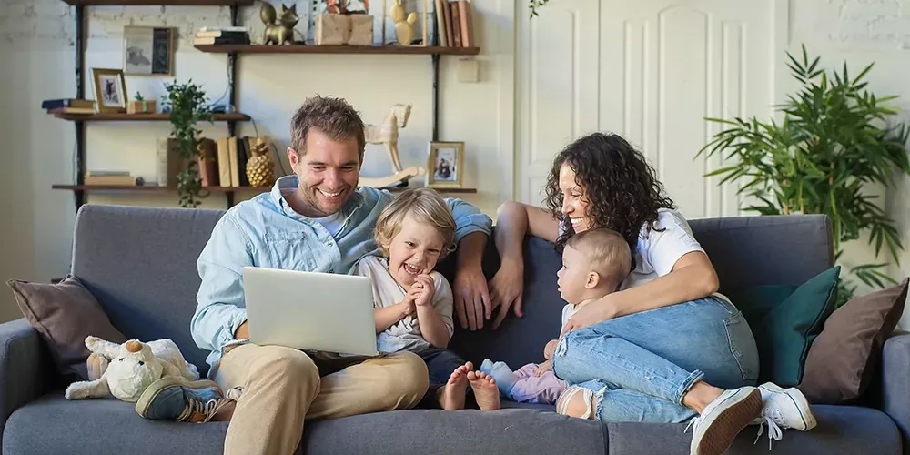 Eine Familie mit zwei kleinen Kindern sitzt zusammen auf einem Sofa. Alle lachen. Der Mann hat einen Laptop auf dem Schoß und scheint seiner Familie dort etwas zu zeigen.