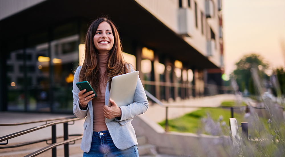 Eine Frau geht lächelnd mit Smartphone und Laptop vor einem modernen Gebäude entlang. Die Szene wirkt urban und geschäftlich.