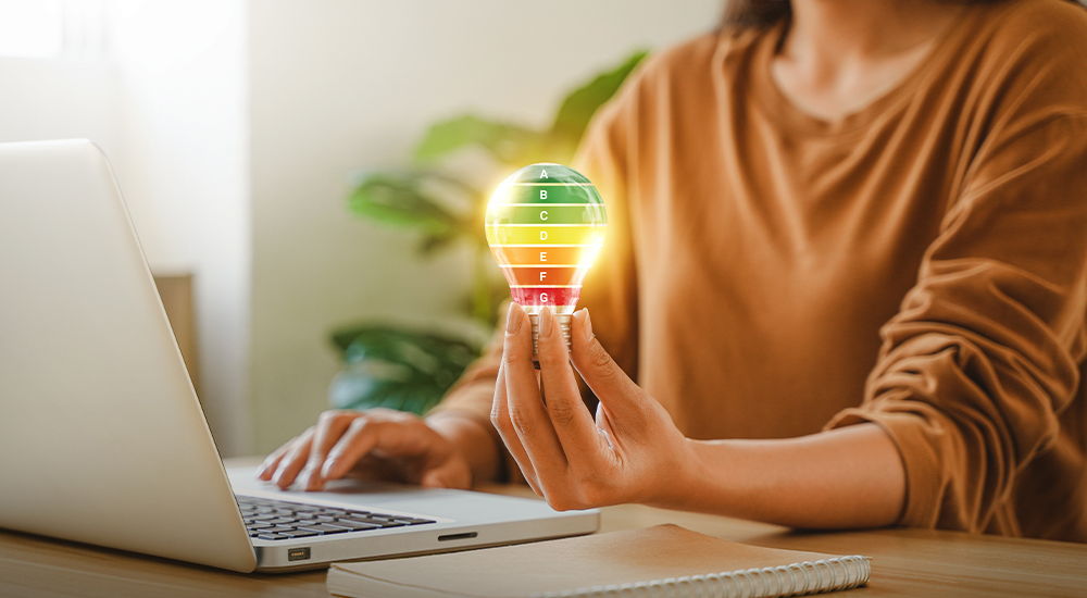 Eine Person hält eine Glühbirne mit Energieeffizienz-Skala neben einem Laptop. Die Szene steht für Energieverbrauch und Effizienz.
