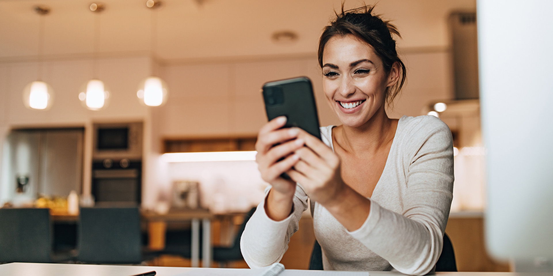Eine Frau sitzt in einer Küche und lächelt, während sie ihr Smartphone benutzt. Die Umgebung ist modern und warm beleuchtet.