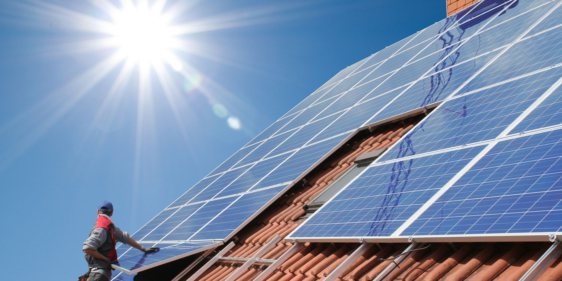 Eine Person installiert Solarpaneele auf einem Hausdach bei sonnigem Wetter. Die Module reflektieren das starke Sonnenlicht.