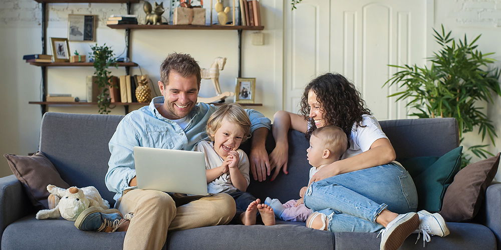 Eine Familie sitzt gemeinsam auf einem Sofa und schaut lachend auf einen Laptop. Das Wohnzimmer ist hell und mit Pflanzen sowie Regalen eingerichtet.